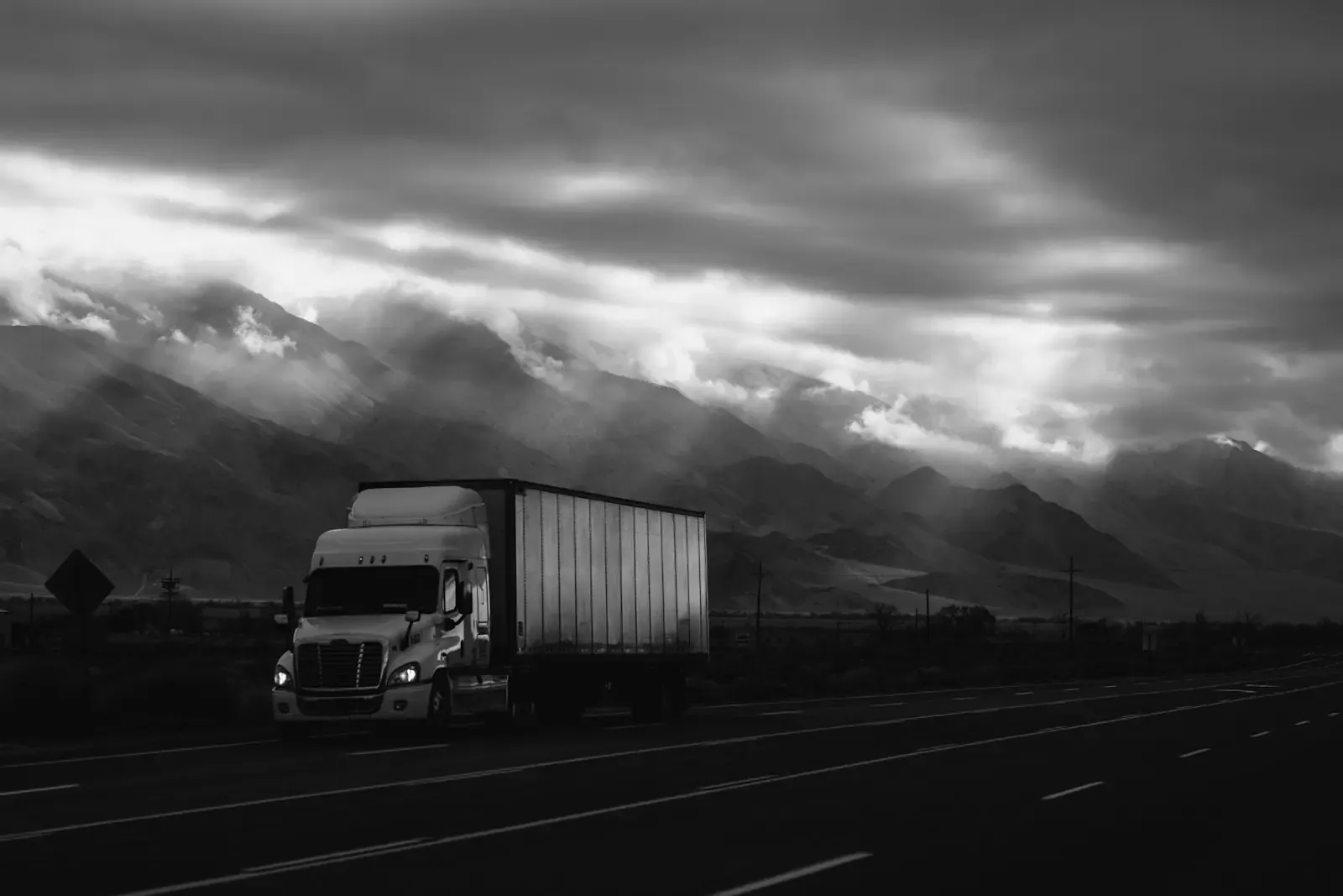 A truck on a highway below cloudy mountains, in black and white